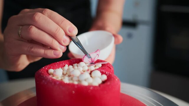 Person carefully decorates a vibrant red cake with white pearls and pink accents. Hands hold a small bowl and tool for precise placement. The cake spins on a turntable. Ideal for food blogs