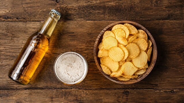 Beer bottle with glass and bowl of potato chips on rustic wooden table. Alcohol drink with salty snack for snack time. Top view of social gathering, party appetizer and pub food.