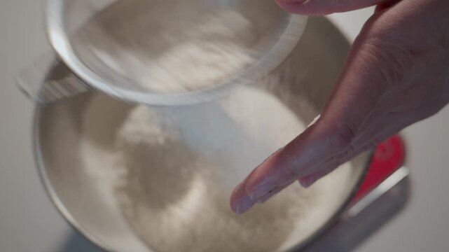 Hand sifts flour into mixing bowl. Close-up shot highlights baking preparation. Ideal for recipes, cooking tutorials, or food blogs