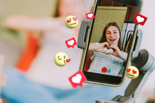 Teenage female influencer showing heart shape while filming through mobile phone with social media icons around at home