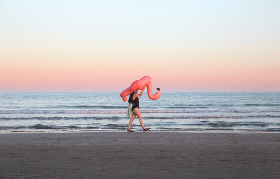 Young woman walking with inflatable pink flamingo on the beach at sunset