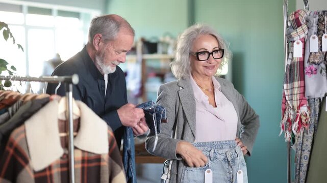 Elderly couple hangs out together in a clothing store and chooses casual jeans with interest. Wife helps retired husband choose denim pants. High quality 4k footage