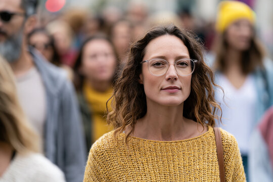 Woman with glasses stands calmly in busy crowd. Her gaze is steady, thoughtful, unbothered by surroundings. Brown curls frame her face, softened by warm tones. Yellow knit sweater adds earthy