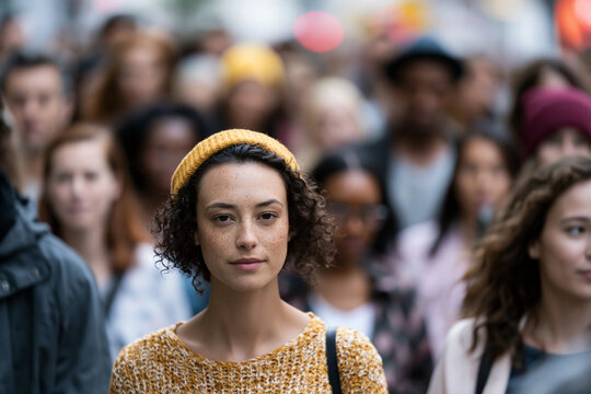 Woman stands out in a bustling crowd. Her gaze holds quiet focus, unbothered by surroundings. Soft, curly hair frames a calm, thoughtful expression. Yellow knitted headband adds a warm, stylish accent