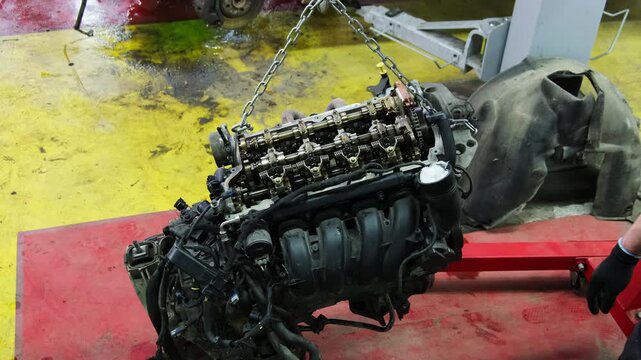 Large car engine hangs on chains in special repair shop. Technician touches surface of engine with hand assessing condition of objects