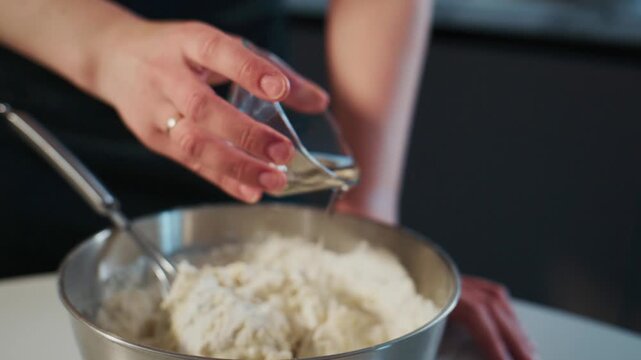 Hand pours liquid into flour mix in metal bowl. Focus on mixing action. Clean kitchen setting. Ideal for baking tutorials or recipe videos. Calm, focused cooking atmosphere