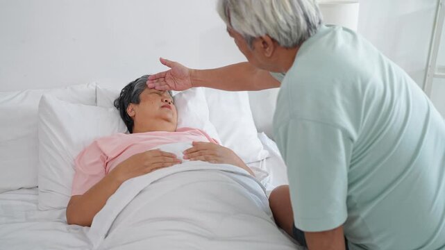Asian senior husband sitting beside wife while resting on bed in house.