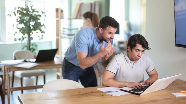 Angry man scolding male colleague working on laptop during stressful workday in office