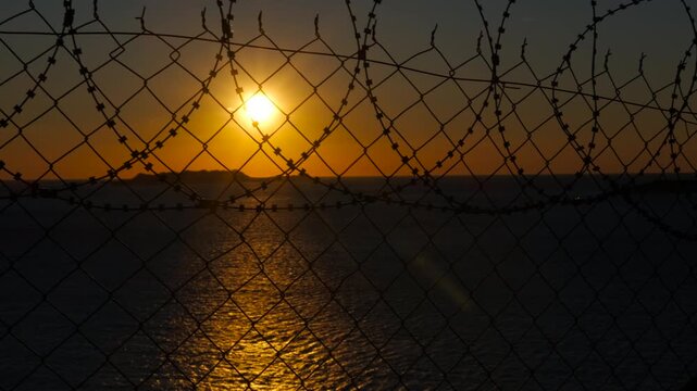 Border at sunset. Orange sun setting over calm body of water, seen through a metal fence topped with barbed wire, creating a poignant contrast between natural beauty and confinement
