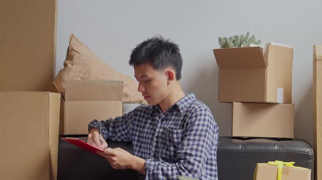 Man checking off items on moving inventory list with cardboard boxes sits on floor in new home