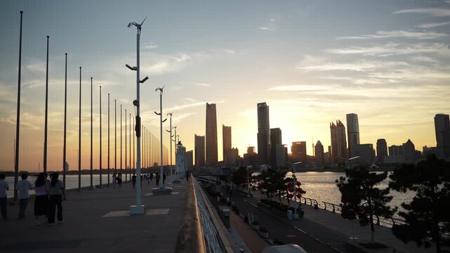 This is a waterfront promenade at dusk, with distant city skyline silhouetted against the warm sunset glow, visitors walking on the paved path.