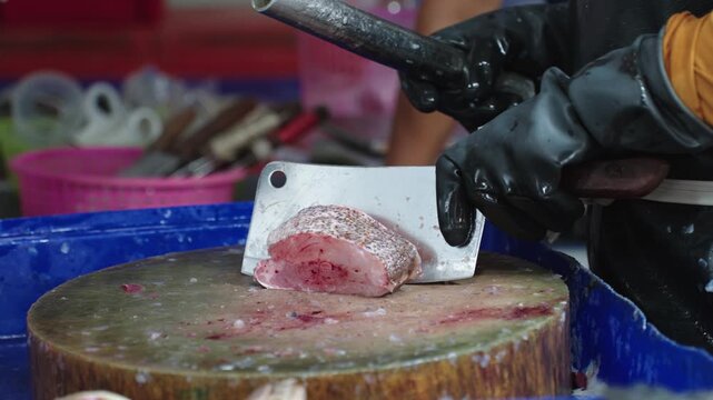 Fishmonger cutting fresh fish steaks with cleaver on wooden chopping block at wet market. Traditional seafood market scene featuring fresh seafood trade in Thailand