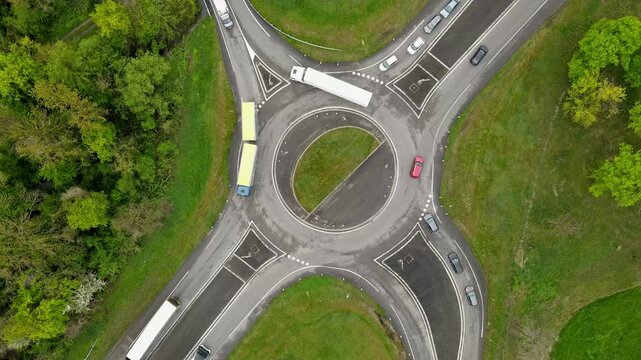 Aerial drone slow zoom-in on roundabout with public transit buses and cars negotiating lanes around green central island. Route geometry and timetable rhythm visible with surrounding green corridor.
