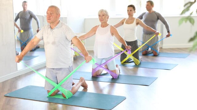 Group of seniors using resistance bands to stretch their arms and body while kneeling on mats. High quality 4k footage
