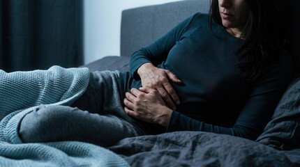 Fototapeta premium Woman Lying in Bed Clutching Her Abdomen Feeling Discomfort in Dark Lighting
