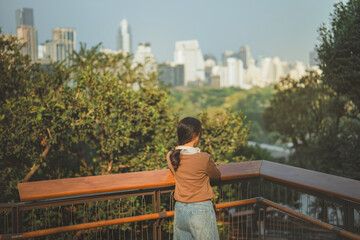Obraz premium Rear view of young Asian woman leaning on wooden balcony rail and looking at city skyscrapers, thoughtful female relaxing at observation deck park in morning, modern urban life, mindfulness and future