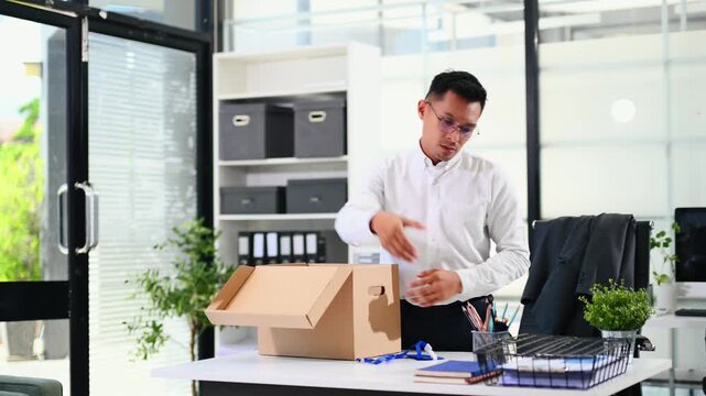 Asian businessman packing personal belongings into a cardboard box, clearing his office desk after resignation