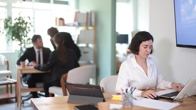 Woman employee of the bank office sits at the table and looks through documents and information on accounts on a laptop. Office worker typing on a laptop against the background of other workers. High