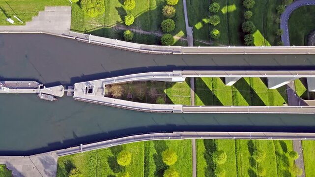 Aerial drone view of vessel navigating narrow canal near boat lift with concrete lock and surrounding green park. Symmetrical waterway with shadow patterns and movement through confined channel.