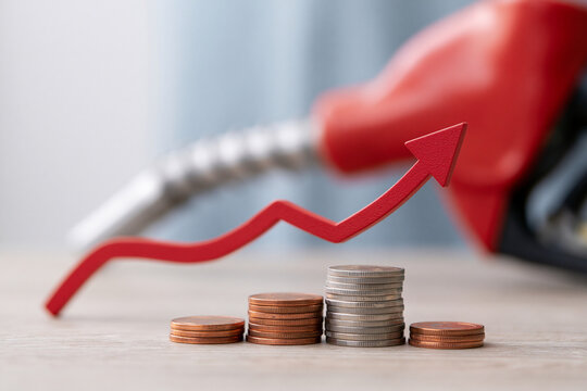 Red arrow rises above coin stacks, signaling rising costs. Fuel pump looms in soft blur behind the scene. Coins stacked low, yet arrow climbs sharply upward
