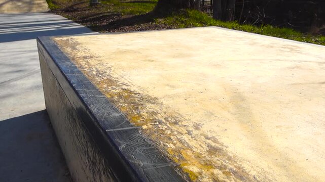 Close up of a well used concrete grind box ledge in an empty urban skatepark, showing wear and wax marks from skateboard trucks sliding along the black metal coping on a sunny day