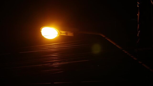 Dramatic low angle night view of tangled electric power cable wires on utility pole with glowing orange street lamp against dark blue sky
