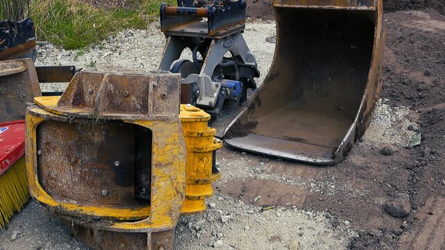 Various attachments for an excavator, including a digging bucket and other parts, resting on gravel and mud at a construction site, ready for heavy earthmoving and building work
