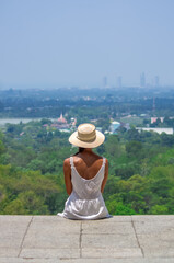 Fototapeta premium Woman in white dress and straw hat sitting on hilltop viewpoint overlooking tropical city landscape in Thailand