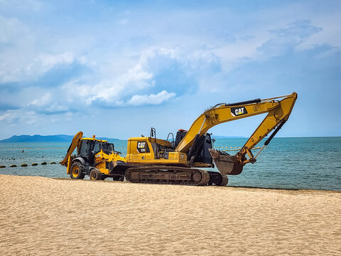Pattaya, Thailand - April 4, 2025: A Caterpillar CAT 320 excavator and JCB loader stand on a sandy beach during coastal construction work with turquoise sea and mountains visible in background.