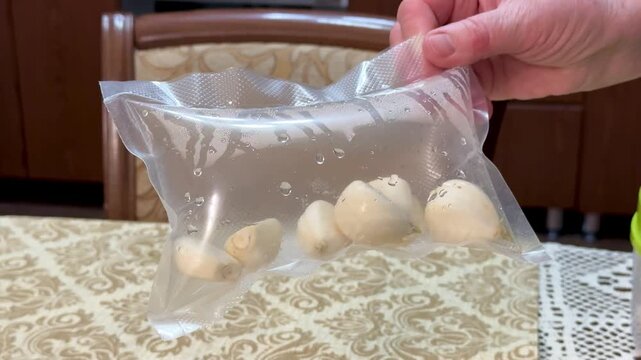 Vacuum bag with garlic cloves on patterned tablecloth, transparent container lifted by hand showing condensation and sealed edge