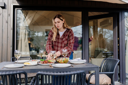 Blonde woman setting outdoor lunch table near modern glass cabin