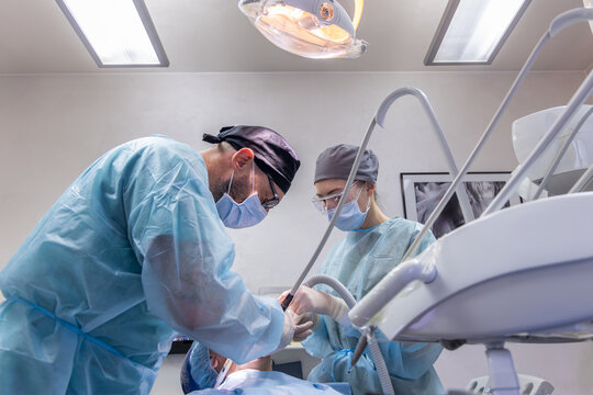 Low angle view of dentist and assistant performing oral surgery in a clinic, with overhead examination light protective gowns suction hose and covered dental unit framing focused teamwork