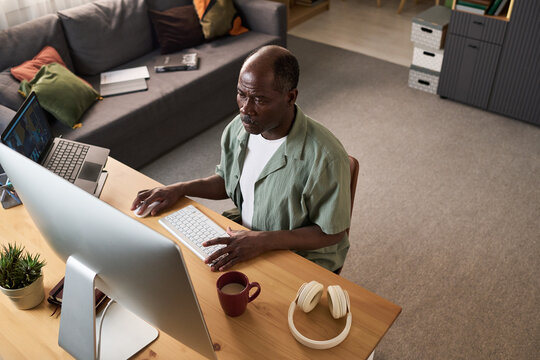 High angle view of African American senior man studying online at home, using desktop computer and laptop for digital upskilling. Ideal for e learning, remote education, reskilling