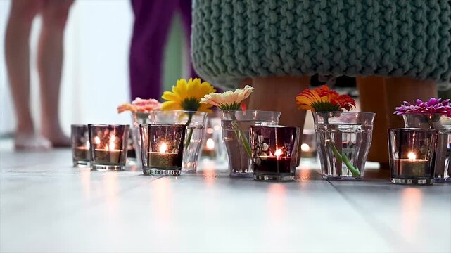 Gentle movement at floor level over glowing tea lights in amber votives alternating with gerbera flowers near turquoise knit pouf. Warm intimate candlelight with soft bokeh in ceremony setting. Floor