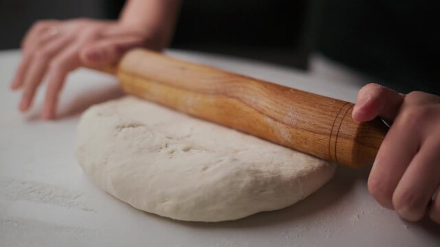 Hands roll dough with wooden roller on white surface. Focus on action, not emotion. Scene shows simple, traditional baking process. Perfect for cooking, recipe, or home kitchen videos
