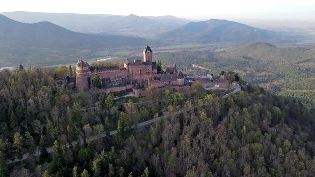 Cinematic aerial drone view of sunset illuminating medieval castle amid rolling Alsace hills with distant village settlements and warm golden atmosphere. Fortress silhouette with battlement outline