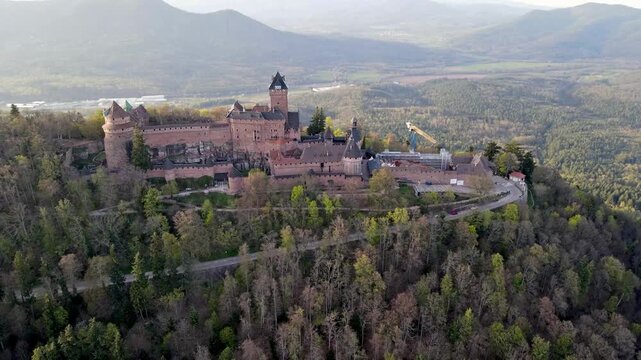 Aerial overview of medieval fortress with stone battlements and tower surrounded by lush green forest and Alsace countryside. Historic defensive structure with ramparts and keep visible from above.
