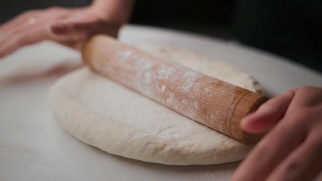 Hands roll dough on white surface. Rolling pin flattens dough slowly. Dusting flour keeps dough from sticking. Close-up shot shows dough texture. Ideal for food, bakery, cooking, or recipe videos