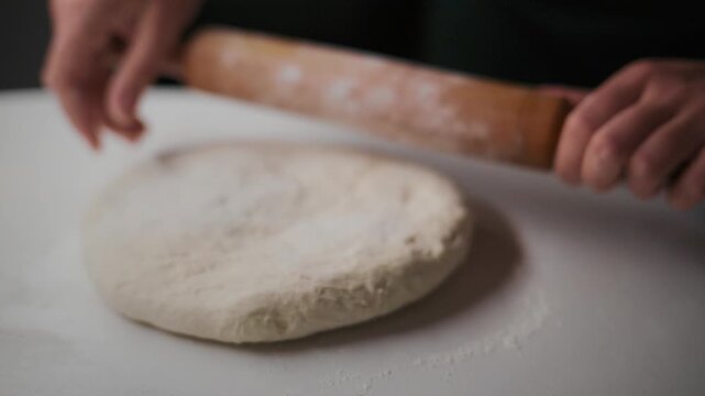 Hands roll dough with wooden roller on white surface. Flour dusts workspace as dough flattens. Process shows careful attention to detail. Ideal for cooking, bakery, or food preparation content