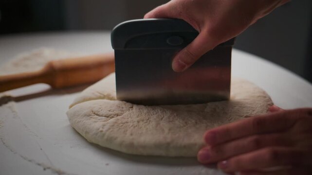 Hand holds a metal dough scraper over risen dough. The tool presses down, cutting the dough cleanly in half. The action is steady, focused, and precise