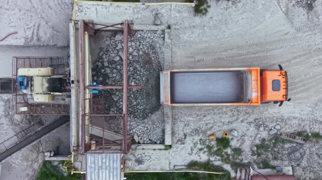A dump truck backs up to a gravel processing plant to unload stones and materials. Workers operate machinery at the site. Sunlight is visible in the scene.