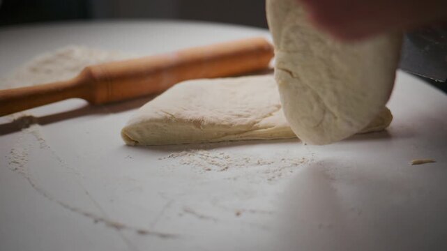 Hand cuts dough with knife on floured surface. Rolling pin rests nearby. Scene shows close-up of pizza preparation process. Ideal for culinary or food-related projects