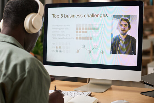African American senior man learning business skills online at home, watching webinar on desktop computer and taking notes. Useful for remote education, upskilling, career training