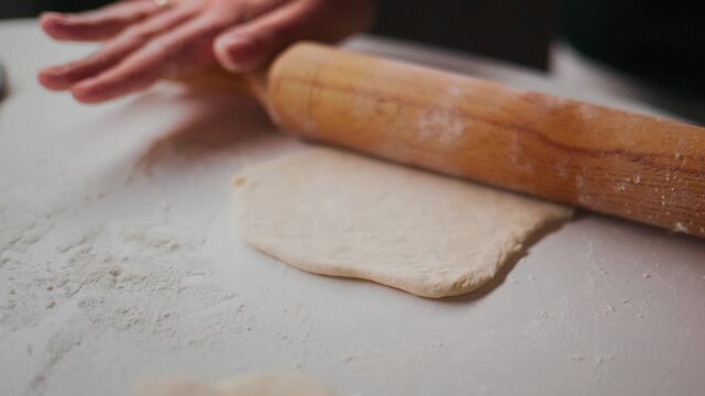 Close-up of hands rolling dough with wooden roller on floured surface. Person focused, careful, precise motion. Ideal for cooking, baking, or food preparation projects. Suitable for recipes, tutorials