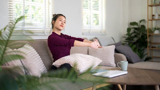 Asian women relaxing stretching arms sitting on sofa in bright living room taking break from work at home feeling peaceful wellness lifestyle healthy living