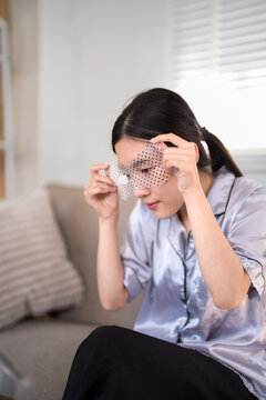 Asian woman applying cooling eye mask gel patch for dark circles puffiness treatment during morning skincare routine relaxing at home on bedroom sofa wearing