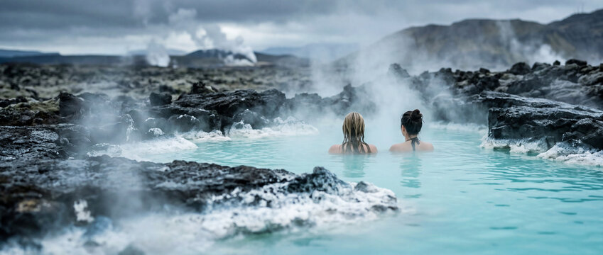 Two women sit shoulder-deep in milky blue geothermal waters, surrounded by jagged volcanic rocks and rising steam, evoking relaxation, travel, nature and wellness in a dramatic landscape.