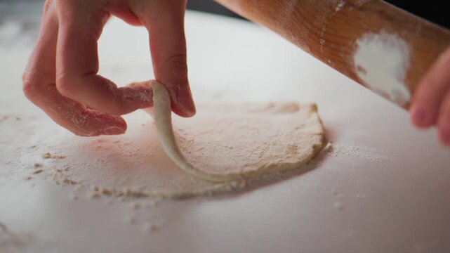 Hands roll dough with wooden roller on floured surface. Flour dusts workspace as dough flattens. Person focuses carefully, showing calm concentration. Ideal for recipes, cooking tutorials