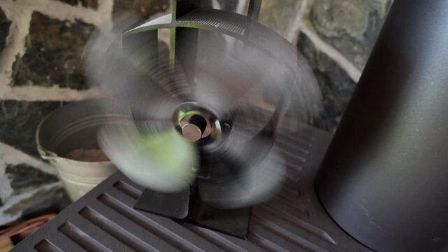 Close‑up of a fast‑rotating stove fan using fire heat for motion, set on a classic cast iron heater with stone background.