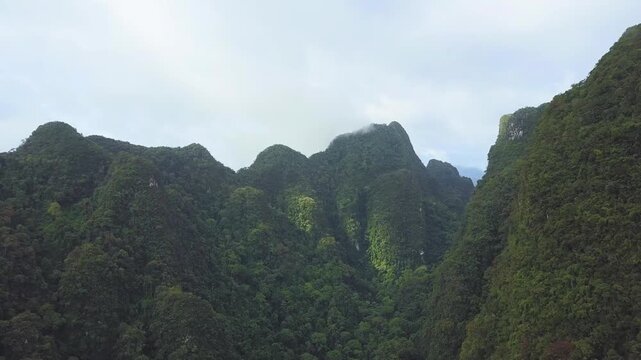 Forward-moving drone shot flying between towering tropical limestone mountains covered in dense rainforest vegetation. Atmospheric aerial footage with dramatic natural formations, misty peaks, and imm